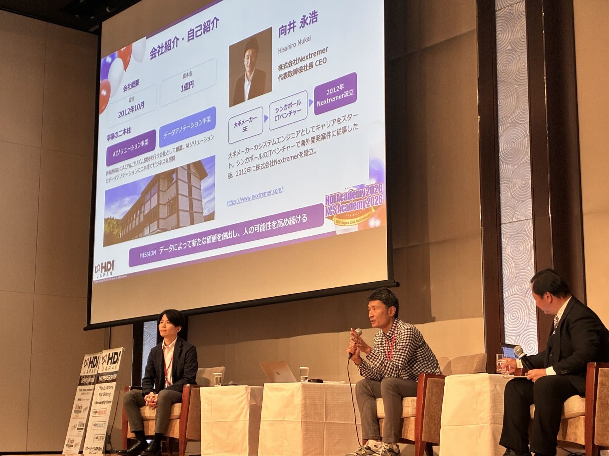 HDIアカデミー2026のセッション「CX向上のためのAI活用」で、自己紹介のスライドを背景にマイクを持って話す株式会社Nextremer 代表取締役CEO 向井永浩